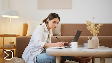 A Czech language student for foreigners is sitting at a laptop and writing handwritten notes for her online Czech lesson.